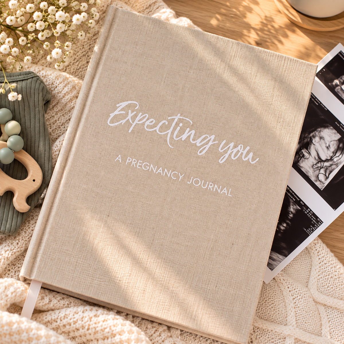 Expecting You pregnancy journal with natural linen cover beside ultrasound photos and a baby teether